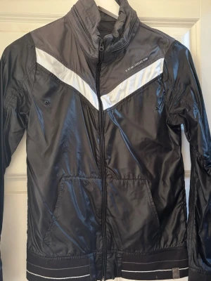 Svart vindjacka från G-Star RAW XS - Snygg svart vindjacka från G-Star RAW i storlek XS. Jackan har vita detaljer i ett V-mönster över bröstet, hög krage och dragkedja framtill. Tillverkad i 100% polyamid med två fickor och elastiska muddar nertill. Perfekt för en sportig och urban stil.