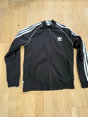 Svart Adidas track jacket - Klassisk svart track jacket från Adidas med vita ränder längs ärmarna och vit Adidas-logga på bröstet. Dragkedja framtill, ribbade muddar och krage. Perfekt för en sportig och avslappnad stil.