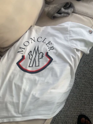 Vit Moncler t-shirt med stor logga - Vit t-shirt från Moncler med stor tryckt logga på framsidan och ett broderat Moncler-märke på ärmen. Klassisk passform och rund halsringning. Perfekt för dig som gillar stilrena och exklusiva plagg.