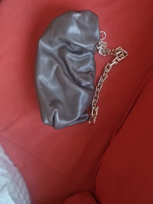 Mörkbrun clutch med kedjehandtag - Trendig mörkbrun handväska i mjukt, glansigt konstläder med en chunky guldfärgad kedja som handtag. Väskan har en puffig, slouchy form och minimalistisk design utan synliga loggor eller detaljer. Perfekt accessoar för en modern look.