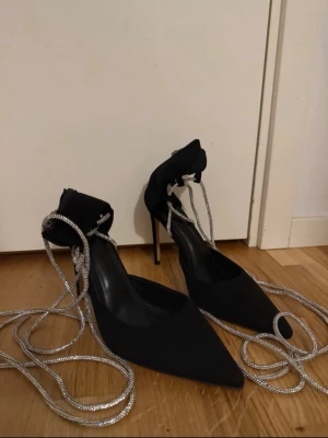Svarta klackskor med strassband och dragkedja  - Snygga svarta klackskor med spetsig tå hög klack  (ca 10-12 cm) och glittrande strassband att snöra runt ankeln. Skorna har dragkedja bak för enkel påtagning och är tillverkade i mockaliknande material. Perfekta för dig som vill sticka ut med detaljer och glam. Skorna har använt bara en enda kväll sedan inget mer. Dock ser ni de smutsiga sulorna, men det är inget som påverkar användningen.