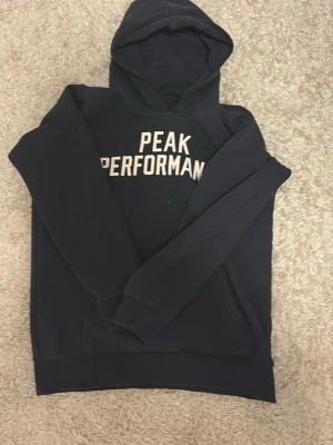Svart hoodie Peak Performance - Säljer en svart hoodie från Peak Performance med vit text på bröstet. Klassisk modell med huva och långa ärmar. Mjuk insida och ribbade muddar vid ärmslut och nederkant. Perfekt för en avslappnad och sportig stil.
