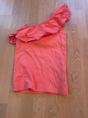 Rosa oneshoulder topp från Molo - Snygg korallrosa oneshoulder topp från Molo med volangdetalj längs axeln. Perfekt för dig som vill sticka ut med en cool och trendig look. Tunn och skön bomullskvalitet som passar bra till sommaren!