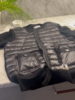 Svart dunväst från Moncler - Snygg svart dunväst från Moncler med quiltad design och två stora fickor med knappstängning. Västen har ribbade detaljer vid krage och nederkant samt dragkedja framtill. Sparsamt använd, och kommer med autenticitet triangeln och moncler prislappen. Skickar också bild på där jag skannar QR koden för att visa autenticitet. Själva moncler lappen inuti jackan har åkt av lite på sidan, men sitter fortfarande fast och går att fixa enkelt genom att använda lite nål och tråd. Kan skicka fler bilder❤️