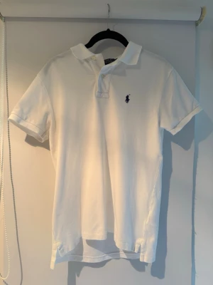 Vit pikétröja från Ralph Lauren - Klassisk vit pikétröja från Ralph Lauren med korta ärmar och blå broderad logga på bröstet. Tröjan har krage och knäppning framtill. Tillverkad i mjuk bomull som känns skön mot huden. Piken är i storleken S i slimfit och är använd max 3 gånger. Bara att skriva vid några funderingar.