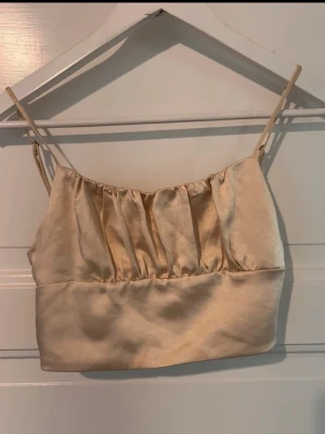 Champagnefärgad satin linnetopp - Snygg champagnefärgad linnetopp i satin med tunna, justerbara axelband och rynkade detaljer framtill och baktill. Perfekt att styla med höga jeans eller kjol för en trendig look.
