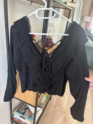 Svart blus med volanger och knytning - Trendig svart blus med långa ärmar, volangdetaljer och knytning framtill. Djup v-ringning med spetskant ger en romantisk vibe. Tillverkad i ett lätt, krinklat tyg som känns luftigt och bekvämt. Perfekt för att lyfta vilken outfit som helst. Använd en gång men behövs strykas.