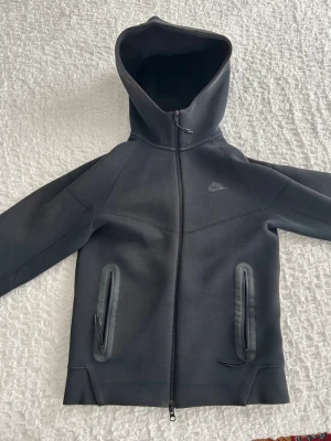 Svart Nike Tech Fleece hoodie - Snygg svart Nike Tech Fleece hoodie med dragkedja framtill och två zip-försedda fickor. Ikonisk Nike-logga på bröstet och modern passform. Tillverkad i mjukt och stretchigt material som ger både stil och komfort. Perfekt för dig som gillar streetwear och sportig look.