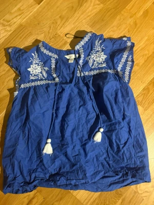 Blå broderad blus med tofsar - Snygg blå blus med vita broderade blommor och dekorativa tofsar framtill. Blusen har korta ärmar och ett avslappnat fall. Perfekt för dig som gillar boho-stil och vill ha något unikt i garderoben.
