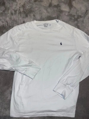 Vit långärmad tröja från Polo Ralph Lauren - Klassisk vit långärmad tröja från Polo Ralph Lauren med den ikoniska blå broderade loggan på bröstet. Tröjan har rund halsringning och är tillverkad i mjuk bomull, perfekt för en clean och stilren look.