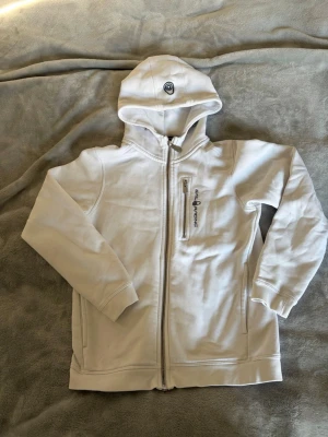 Vit hoodie från Sailracing - Snygg vit hoodie från Sailracing med dragkedja framtill och en praktisk bröstficka med logga. Hoodien har en broderad detalj på huvan och ribbade muddar vid ärmar och nederkant. Perfekt för en avslappnad och sportig stil. Det finns fläckar på luvan som ej går bort i tvätten, därav det låga priset. 