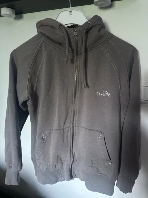 Grå zip-hoodie från Oxide - Säljer en grå hoodie från Oxide med dragkedja framtill och två stora fickor. Tröjan har huva med dragsko och vita kontrastsömmar. Perfekt för chill dagar och enkel att matcha med jeans eller joggers.