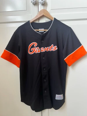Svart Giants baseballtröja - Snygg svart baseballtröja med Giants-logga i orange och vitt på bröstet. Tröjan har korta ärmar med breda orangea muddar och vita detaljer. Knäppning framtill med svarta knappar. Perfekt för sport eller streetwear.