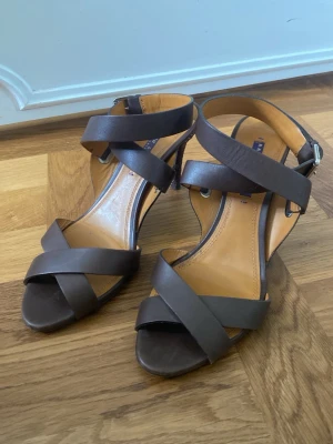 Bruna pumps med remmar och klack - Snygga bruna pumps från Ralph Lauren i läder med remmar över foten och runt ankeln. Klack med ormmönstrade detaljer. Står 9B under skon, själv har jag 39 och dom passar perfekt. Skon har används endast ett fåtal gånger (därav slutningarna under skon som syns på sista bilden), i övrigt jättebra skick. 