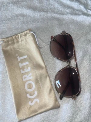 Solglasögon från Scorett - Snygga solglasögon från Scorett med klassisk pilotform och transparent brunmelerad båge. Glasen är mörktonade i brunt och ger en cool vibe till din look. Kommer med en beige Scorett-påse för förvaring. Aldrig Använda!