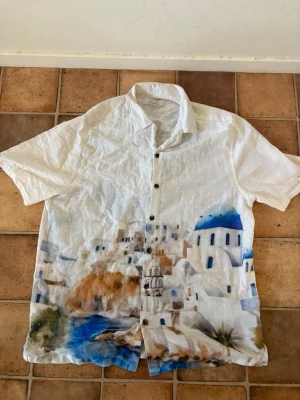 Vit linneskjorta med Santorini-motiv. köpte i Tyskland  - Vit kortärmad linneskjorta från Mondo med färgglatt tryck av Santorini på framsidan. Skjortan har knappar framtill och avslappnad passform, perfekt för varma dagar. Motiv i blått, brunt och grönt ger en chill vibe.