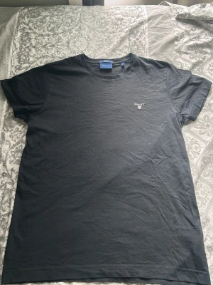Svart t-shirt från GANT - (FRÅN EN HOCKEYSPELARE I ÖREBRO) Klassisk svart t-shirt från GANT med broderad logga på bröstet. T-shirten har rund halsringning och korta ärmar. Tillverkad i mjuk bomull som känns skön mot huden. Perfekt basplagg för garderoben.