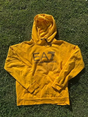 Gul EA7 Emporio Armani hoodie - Snygg gul hoodie från EA7 Emporio Armani med stor logga på bröstet. Hoodien har huva med dragsko och en klassisk känguruficka framtill. Tillverkad i mjukt bomullsmaterial och har ribbade muddar vid ärmslut och nederkant. Perfekt för en avslappnad och trendig look.