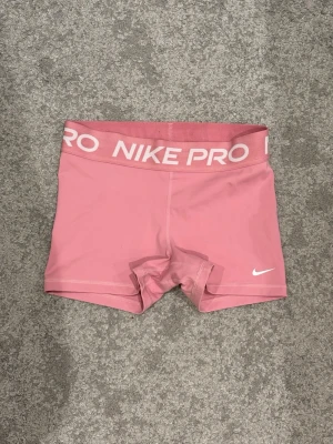 Rosa Nike Pro shorts - Säljer ett par rosa Nike Pro shorts med bred resår i midjan där det står 'NIKE PRO' i vitt. De har en tight passform och är perfekta för träning. Materialet är stretchigt och känns bekvämt mot huden. Liten vit Nike-logga på benet.