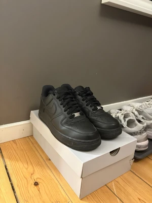 Nike Air Force 1 helsvarta  - Snygga helsvarta Nike Air Force 1 sneakers i klassisk modell. Skorna är i läder med perforerad tåbox, svart sula och svarta skosnören. Perfekta för dig som gillar en clean och tidlös stil. Ikonisk Nike Swoosh på sidorna och metallbricka på snörningen.