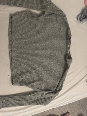 Grå ribbad stickad tröja - Mysig grå stickad tröja med ribbad struktur och rund hals. Tröjan har lång ärm och en enkel, rak passform som gör den lätt att matcha med olika outfits. Perfekt för lager-på-lager och chill dagar.
