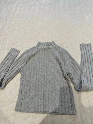 Grå ribbstickad polotröja - Säljer en grå ribbstickad polotröja med lång ärm och hög hals. Tröjan har en smal passform och är tillverkad i mjukt material som passar perfekt till kyligare dagar. Enkel och stilren design som är lätt att matcha.