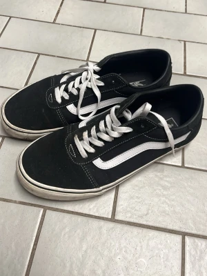 Svarta Vans Old Skool sneakers - Klassiska Vans Old Skool sneakers i svart med vit sula och vit sidostripes. Skorna har vita skosnören och ovandel i mocka och canvas. Perfekta för en avslappnad streetwear-look och passar till det mesta i garderoben.