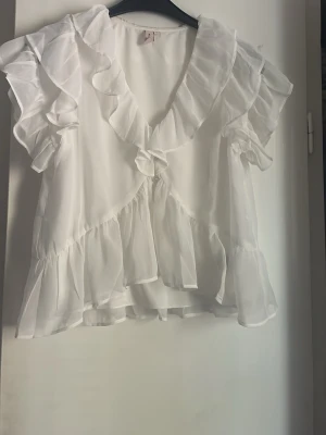 Vit volangblus med kort ärm - Söt vit blus i transparent, lätt material med volangdetaljer och v-ringning. Blusen har korta ärmar och flera lager volanger.