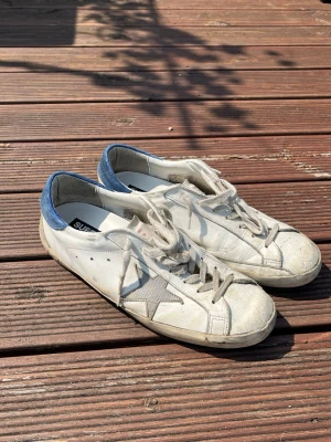 Golden Goose Superstar   - Snygga vita sneakers från Golden Goose med grå stjärndetalj på sidan och blå mockadetalj vid hälen. Skorna har vita snören och blå sula undertill. Perfekta för en avslappnad streetstyle-look.