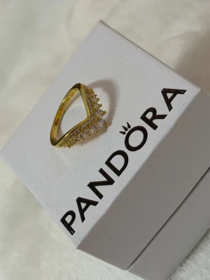 Guldfärgad ring med stenar från Pandora - Snygg guldfärgad ring från Pandora i storlek 54. Ringen har en V-formad design och är dekorerad med gnistrande klara stenar längs kanten som ger en lyxig känsla. Perfekt för dig som vill ha något extra till din stil.