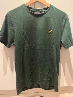 Grön t-shirt från Lyle & Scott - Mörkgrön t-shirt från Lyle & Scott med klassisk passform och korta ärmar. T-shirten har rund halsringning och den ikoniska gula örnloggan broderad på bröstet. Tillverkad i mjuk bomull för en skön känsla.