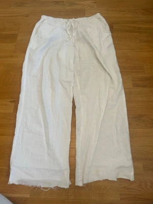 Vita linnebyxor från Gina Tricot - Säljer ett par vita linnebyxor från Gina Tricot med avslappnad passform och resår i midjan samt dragsko. Byxorna har raka ben och är tillverkade i ett luftigt linnematerial, perfekta för varma dagar. Klippta längst ner vid benen 