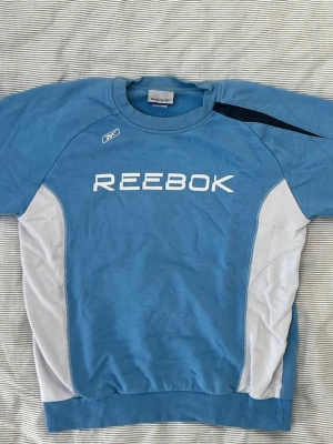 Vintage Reebok blå och vit sweatshirt - Blå sweatshirt från Reebok med vit logga framtill och vita paneler på sidorna. Tröjan har rund halsringning, långa ärmar och en svart detalj på axeln. Tillverkad i mjukt material, perfekt för träning eller chill.