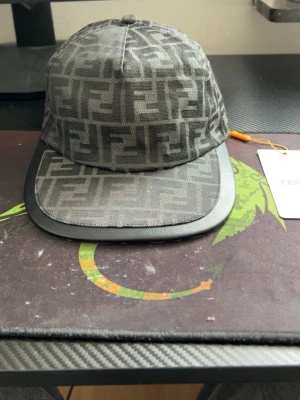 Svart keps F - Snygg svart keps från F med det klassiska FF-monogrammet över hela kepsen. Kepsen har böjd skärm med läderdetalj och justerbar baksida. Perfekt för dig som vill ha en exklusiv och trendig accessoar med tydlig logga.