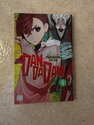 Dandadan Volym 1 av Yukinobu Tatsu - Manga-bok 'Dandadan' volym 1 av Yukinobu Tatsu. Omslaget har färgstark illustration med en tjej med rött hår och stora örhängen samt en mystisk figur i bakgrunden. Boken är på engelska och tryckt i mjukt omslag. Perfekt för dig som gillar science fiction och övernaturliga äventyr.