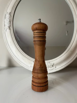 Klassisk pepparkvarn i trä - Säljer en stilren pepparkvarn i brunt trä med svarvade detaljer och metallknopp på toppen. Kvarnen har en elegant, avsmalnande form och svarta dekorlinjer. Perfekt för att mala peppar direkt över maten.