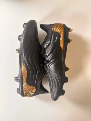 Adidas Copa Sense svart/guld fotbollsskor - Snygga Adidas Copa Sense fotbollsskor i svart med guldiga detaljer på hälen och sulan. Skorna har en modern, vågig design och är tillverkade i syntetmaterial med snörning för bra passform. Perfekta för dig som vill sticka ut på planen. (Hjärndobbar) 39 och en halv fast passar 40-41