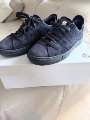 Mörkblå Moncler sneakers i mocka - Snygga mörkblå sneakers från Moncler i mjuk mocka med matchande mörkblå sula och snörning. Klassisk låg modell med diskret Moncler-logga på plösen. Perfekta för dig som gillar stilrena och exklusiva skor.