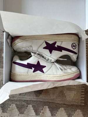 Bape Sta vita sneakers med lila stjärna - Säljer ett par vita Bape Sta sneakers med ikonisk lila stjärndesign på sidan och matchande lila sula. Skorna har klassisk låg profil, perforerad tåbox och vita skosnören. Tillverkade i läder för en clean och stilren look.