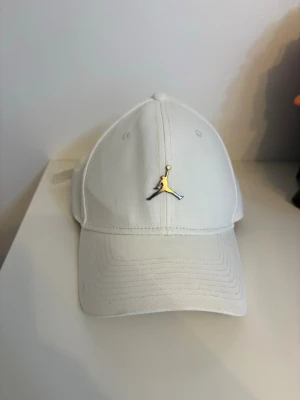 Vit Jordan keps  - Snygg vit keps från Jordan med den ikoniska Jumpman-loggan i guld framtill. Klassisk böjd skärm och justerbart spänne bak för perfekt passform. Tillverkad i slitstarkt bomullstyg som ger en clean och sportig look.