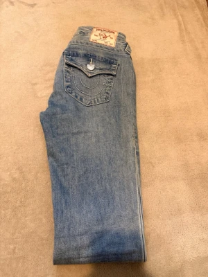 True Religion ljusblå jeans - Säljer ett par ljusblå jeans från True Religion med deras ikoniska bakficka och kontrastsömmar. Jeansen har en klassisk femficksmodell och är tillverkade i mjukt denimtyg. Perfekta för en avslappnad och trendig look.