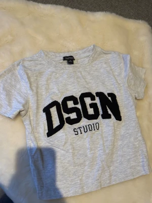 Grå t-shirt DSGN Studio Boohoo - Snygg ljusgrå tight t-shirt från Boohoo med stor svart text 'DSGN Studio' framtill. Klassisk passform med korta ärmar och rund hals. Perfekt till jeans eller shorts för en avslappnad look.
