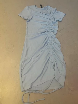 Ljusblå ribbad klänning H&M Divided - Kort ribbad klänning från H&M Divided i ljusblått. Klänningen har korta ärmar, rund halsringning och snörning längs sidan som ger en snygg rynkeffekt. Perfekt för sommarens alla tillfällen! Har inte använt jättemycket men är i bra skick 