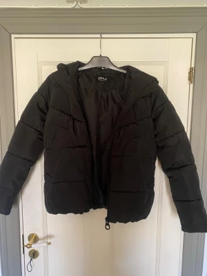 Svart pufferjacka från ONLY - Snygg svart pufferjacka från ONLY med huva och dragkedja framtill. Jackan har en klassisk quiltad design och är vadderad för extra värme. Perfekt för kalla vinterdagar och enkel att matcha med olika outfits.