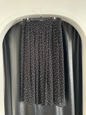 Svart prickig långkjol med resårmidja - Säljer en svart långkjol med vita prickar och bred resår i midjan. Kjolen har ett flowy fall och är perfekt för dig som gillar en avslappnad men ändå stilren look. Materialet känns lätt och skönt mot huden.
