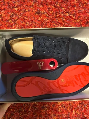 Svarta sneakers från Christian Louboutin - Snygga svarta sneakers från Christian Louboutin med klassisk röd sula och diskret logga. Skorna har en låg silhuett, snörning och är tillverkade i mocka och läder. Perfekta för dig som vill ha en exklusiv och stilren look.