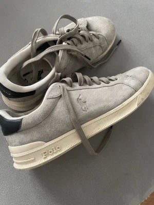Gråa Polo Ralph Lauren sneakers - Snygga gråa sneakers från Polo Ralph Lauren i mocka med klassisk låg profil. Skorna har vita sulor, gråa skosnören och svart detalj vid hälen. Polo-loggan syns på sidan och hälen. Perfekta för en clean och stilren look.