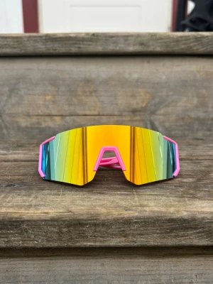 Barnstorlek Sportiga solglasögon med spegelglas - Sportglasögon i barnstorlek, kan också passa äldre med litet ansikte. Bågarna är rosa och har en futuristisk, kantig form som sticker ut. Perfekta för dig som vill ha en sportig vibe och synas i mängden. Skiftande glas i orange, grön och blå. Unisex. 