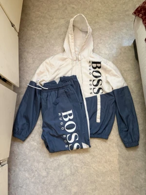 Hugo Boss blåvit tracksuit set - Snyggt tracksuit set från Hugo Boss i blått och vitt. Setet består av en hoodie med dragkedja och stora logotyper samt matchande byxor med resår i midjan och logga längs benet. Perfekt för sport eller chill. Materialet är lätt och andas. Lite smutsigt men syns inte från när eller långt håll men material som en regnjacka!