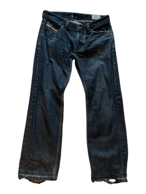 Blåa straight leg jeans från Diesel - Säljer ett par mörkblå straight leg jeans från Diesel med klassiska fem fickor och slitna detaljer vid bensluten. Jeansen har låg midja och coola sömmar på bakfickorna. Perfekta för dig som gillar en avslappnad och trendig stil.